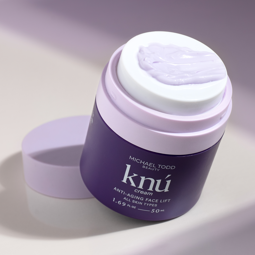 White .  An open jar of Michael Todd Beauty 4 in 1 Dermaplaning & Post Treatment System with visible purple cream, set against a soft-focus lilac background.