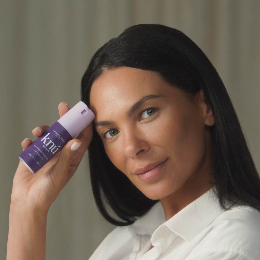 A woman holds a bottle of Knú Serum (30 day) by Michael Todd Beauty, promoting skin regeneration.