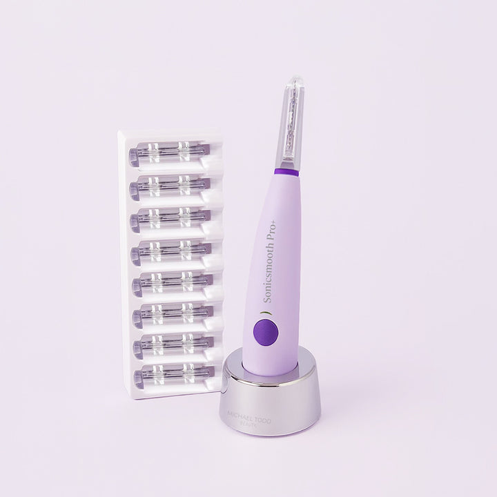 A purple Michael Todd Beauty Sonicsmooth Basic device stands on a charging base beside a tray with eight replacement heads, all set against a light background.